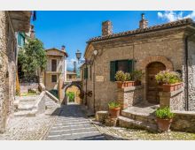 Stradina in pietra lastricata in un borgo storico, con scalinate e case in muratura dai tetti in coppi e persiane verdi. In primo piano si vedono un portale ad arco, un lampione e terrazze con vasi di fiori e piante lungo i muri.