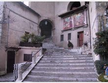 Scalinata in pietra e mattoni che sale in un cortile storico, con archi in muratura e facciate intonacate. Sulla parete a destra sono visibili un grande affresco, una porta in legno e piccole finestre con inferriate, con piante e decorazioni lungo i lati.