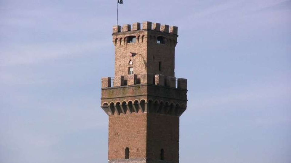 Torre in mattoni in stile medievale con sommità merlata e coronamento aggettante sostenuto da archetti, con piccole feritoie e finestre. La torre si staglia isolata contro un cielo azzurro.