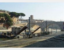 Veduta di un ponte storico in pietra ad arcate su un fiume, con una banchina pavimentata in primo piano e una struttura metallica con rampe d’accesso lungo la riva. Sullo sfondo si vedono edifici e alberi sul lungofiume.