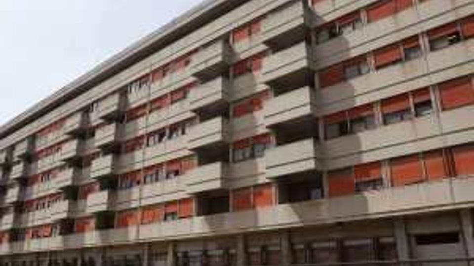 Edificio residenziale moderno a più piani, lungo e lineare, con file di balconi e pannelli/fasce rosse sulle facciate. Al livello strada si vede un porticato continuo e una recinzione metallica lungo il marciapiede.