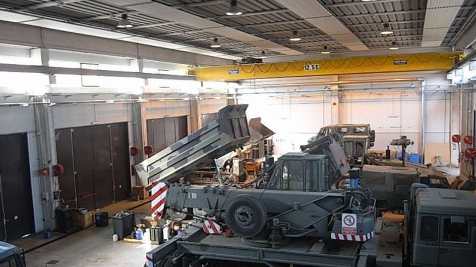 Interno di un grande capannone industriale adibito a officina, con tetto in lamiera e illuminazione a sospensione. Sul pavimento sono presenti vari veicoli militari e camion in manutenzione, attrezzature da lavoro e un carroponte giallo che attraversa la struttura.