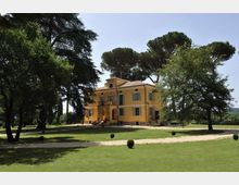 Villa storica color ocra a due piani con tetto in coppi e persiane, al centro di un ampio parco. Davanti all’edificio si vedono prati curati, vialetti in ghiaia e grandi alberi ad alto fusto che incorniciano la facciata.