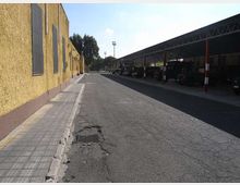 Cortile asfaltato tra un lungo edificio giallo con finestre strette e un’ampia tettoia metallica con pilastri, simile a un deposito o autorimessa. Sulla strada si notano crepe e una griglia di scolo, con alberi e lampioni sullo sfondo.