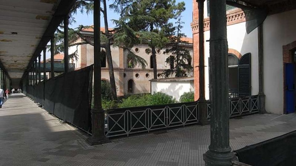 Portico lungo con pavimento piastrellato e tettoia sorretta da colonne metalliche, affiancato da una recinzione bassa. Sullo sfondo si vede un cortile con alberi alti e un edificio in muratura e intonaco con archi e dettagli in mattoni.