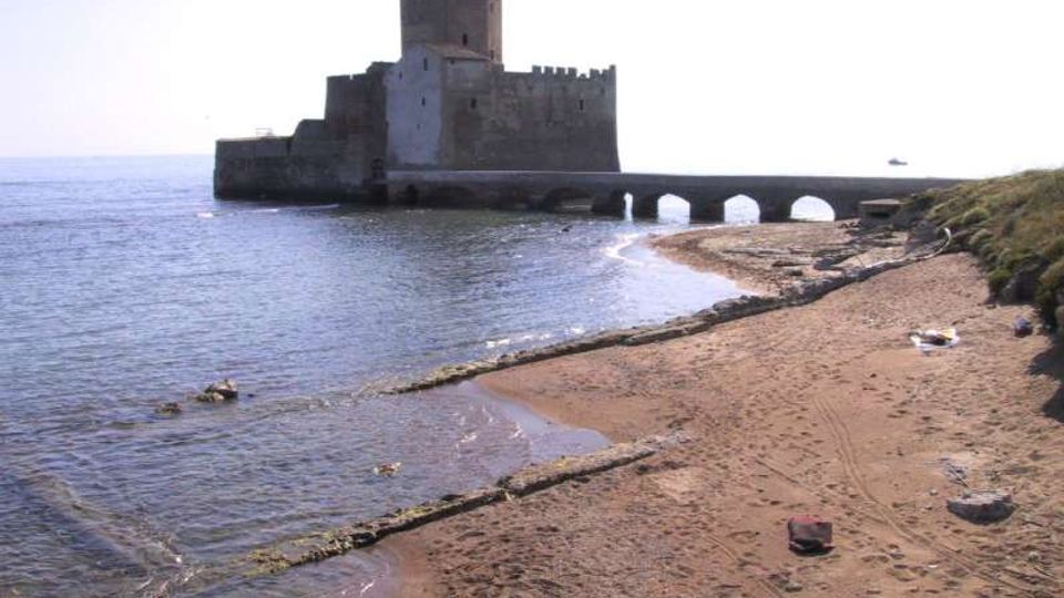Castello costiero in pietra su un isolotto, collegato alla riva da un ponte basso con arcate, affacciato sul mare calmo. In primo piano si vede una spiaggia sabbiosa con scogliera e vegetazione bassa lungo il margine.