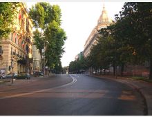 Ampio viale urbano curvo con doppia linea centrale, fiancheggiato da alberi e marciapiedi. Ai lati si vedono palazzi storici con facciate ornate e, sullo sfondo, un grande edificio con cupola.