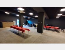 Ampio spazio interno tipo sala eventi con pareti scure e pavimento in cemento, illuminato da pannelli a soffitto e con colonne centrali. Sono presenti tavoli rettangolari coperti da tovaglie rosse con sedie, un bancone in legno su un lato e una zona lounge con divani vicino alle finestre.