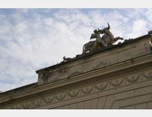 Facciata superiore di un edificio in stile classico con cornici e fregi decorativi in pietra, ripresa dal basso. Sul tetto è visibile un gruppo scultoreo con cavallo e figure, stagliato contro un cielo parzialmente nuvoloso.
