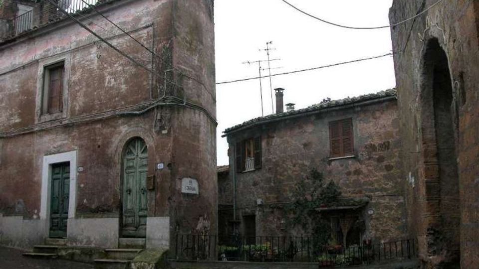 Piccola piazza di borgo con edifici in muratura intonacata e facciate consumate, porte e persiane in legno e scalini in pietra. In primo piano si vedono una ringhiera bassa, cavi aerei e un grande arco in pietra sul lato destro.