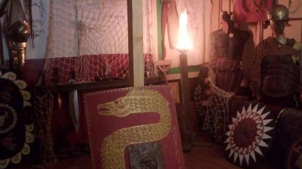 Interno di una piccola sala espositiva con scudi decorati, armature e oggetti storici disposti lungo le pareti. Sono visibili reti appese, pannelli dipinti e una luce centrale che illumina l’ambiente.