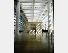 Ampio atrio museale moderno con colonne cilindriche bianche, pavimento lucido e grande soffitto a griglia con lucernari che lasciano entrare luce naturale. Al centro è esposto un monumento in pietra con bassorilievi e una scalinata, circondato da vetrate su entrambi i lati.