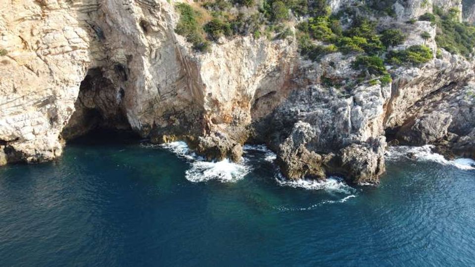 Scogliera rocciosa a picco sul mare con una grande grotta naturale alla base e speroni di roccia frastagliati lambiti dalle onde. Acqua blu scura in primo piano e vegetazione rada sulla sommità della parete calcarea.