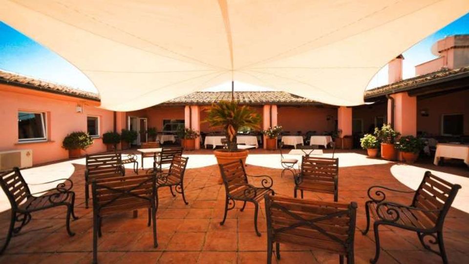 Cortile interno con pavimentazione in terracotta, sedute in legno e metallo disposte al centro e un grande telo bianco teso come copertura ombreggiante. Intorno si vedono edifici bassi color rosa con tetti in coppi e piante in vaso lungo il perimetro.