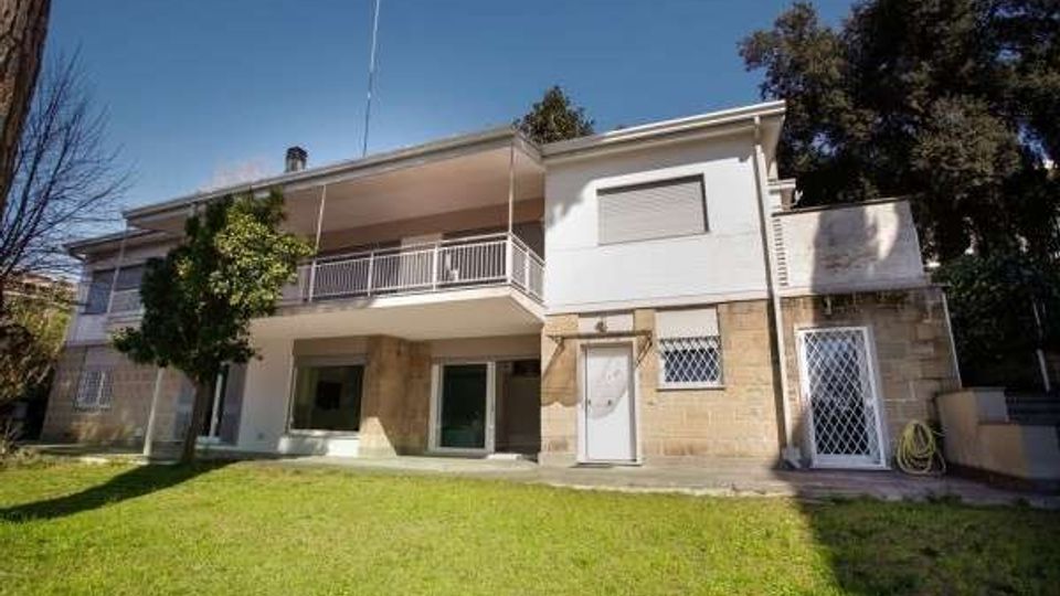 Casa unifamiliare a due piani con facciata in pietra e intonaco bianco, dotata di grande balcone con ringhiera e finestre con persiane. Davanti si estende un prato verde con un albero, circondato da vegetazione e alberi alti sullo sfondo.