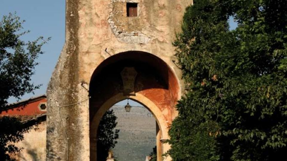 Antica porta d’accesso in pietra con torre merlata e grande arco che sovrasta una strada, con una piccola finestra e segni di intonaco consumato. Alberi e vegetazione fiancheggiano l’arco e la carreggiata, con colline visibili sullo sfondo.