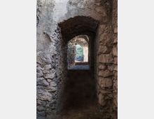 Corridoio stretto in pietra grezza con soffitto a volta e una serie di archi che conducono verso un’apertura luminosa. In fondo si intravede l’esterno con vegetazione, mentre il pavimento è in terra o pietra scura.