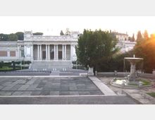 Veduta di un grande edificio monumentale in stile neoclassico con ampia scalinata centrale e portico colonnato. In primo piano c’è una piazza lastricata e, sulla destra, una fontana circolare, con alberi e giardini ai lati.