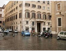Facciata di un palazzo storico in pietra su una piazza lastricata in sampietrini, con finestre ad arco, persiane scure e un portale centrale. Davanti all’edificio si vedono auto e motocicli parcheggiati, dissuasori sferici e una strada urbana tra altri edifici in muratura.