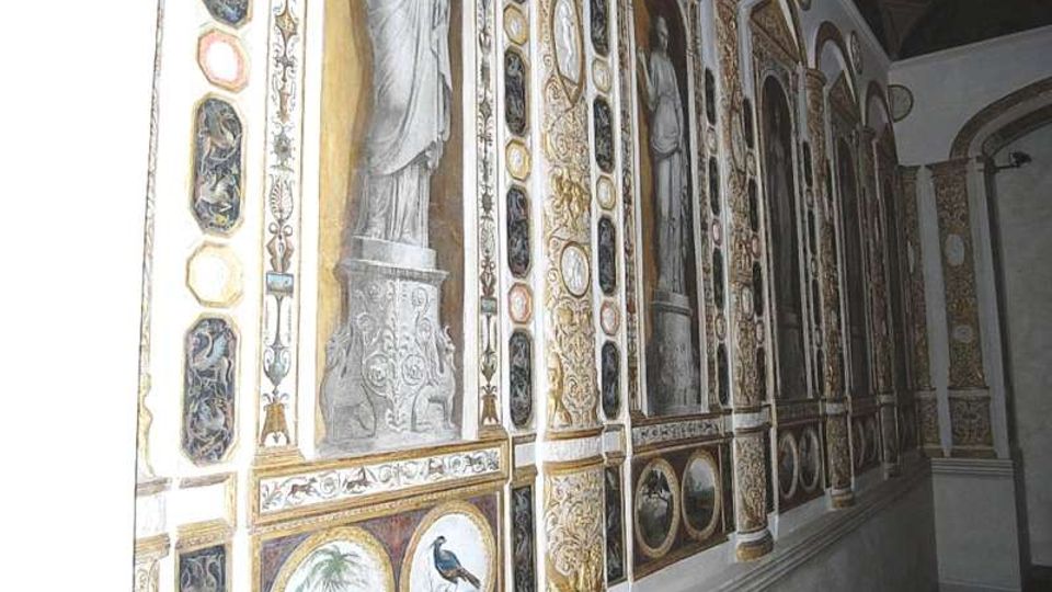 Parete interna di un edificio storico decorata con affreschi e stucchi dorati, con nicchie ad arco contenenti figure monocrome e pannelli ovali con paesaggi e uccelli. Un corridoio stretto corre lungo la parete, con luce intensa proveniente da sinistra.