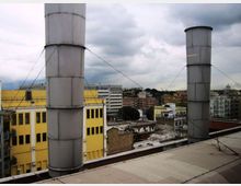 Tetto di un edificio con due alte canne fumarie cilindriche in metallo, sostenute da tiranti, affacciato su una zona urbana con palazzi e magazzini. Sullo sfondo si vedono edifici multipiano e un cielo nuvoloso.