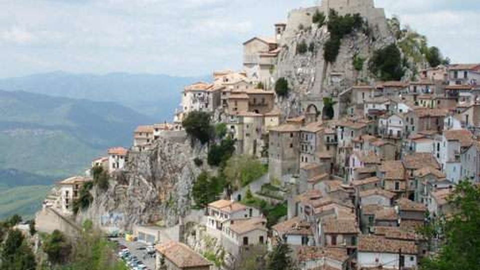 Borgo storico arroccato su uno sperone roccioso, con case in pietra dai tetti in coppi addossate lungo il pendio e una fortificazione in cima. Sullo sfondo si vedono colline e montagne, con una strada e alcune auto ai piedi della rupe.