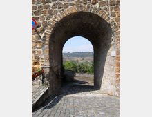 Arco in pietra di una porta medievale con pavimentazione in selciato e mura laterali, che incornicia la vista su una valle con campi e colline in lontananza. Sul lato sinistro si vedono una fioriera e un corrimano lungo la rampa in discesa.