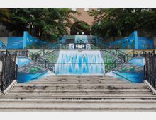 Scalinata urbana in cemento con corrimano metallici, fiancheggiata da grandi murales azzurri che raffigurano una cascata, acqua e rocce. In cima si vedono alberi e la facciata di un edificio in mattoni sullo sfondo.