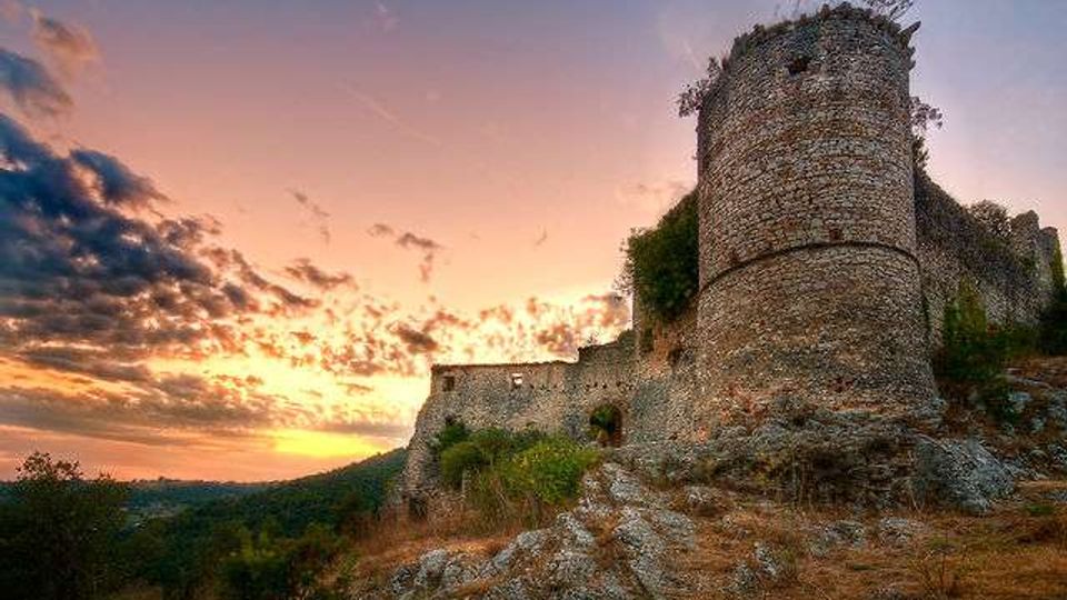 Rovine di un castello medievale su una collina rocciosa, con una grande torre cilindrica in pietra in primo piano e tratti di mura sullo sfondo. Il paesaggio circostante è collinare e boscoso, illuminato da un cielo al tramonto con nuvole sparse.