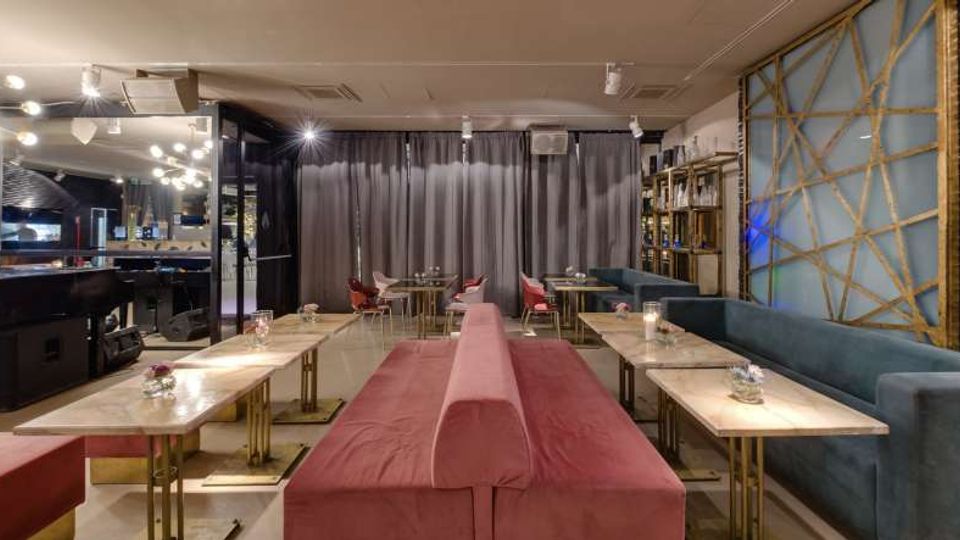 Interno di un lounge bar moderno con lunga panca centrale rivestita in tessuto rosso e tavolini bassi in marmo con basi metalliche. Lungo la parete destra ci sono divanetti blu e una parete decorativa con pannelli illuminati, mentre sul fondo si vedono tende grigie e illuminazione a faretti sul soffitto.