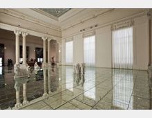 Ampia sala interna in stile neoclassico con colonne e cornici ornamentali, grandi finestre con tende bianche e soffitto alto. Il pavimento lucido a specchio riflette l’architettura e alcune sculture disposte nello spazio.