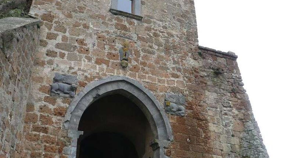 Portale d’ingresso in pietra di una struttura storica fortificata, con grande arco a tutto sesto che introduce a un passaggio coperto. Sopra l’arco si vede una torretta in muratura con tre arcate aperte e un piccolo finestrino, circondata da muri in pietra e pavimentazione in ciottoli.
