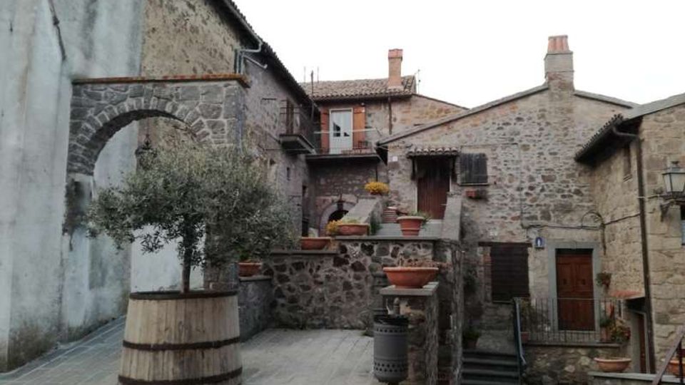 Piccola piazza in un borgo con case in pietra, archi e scale che collegano livelli diversi. Al centro ci sono grandi fioriere e un ulivo in una botte, con muri in pietra e porte in legno sullo sfondo.