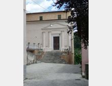 Facciata di una chiesa su una piccola strada in salita, con scalinata centrale in pietra che conduce a un portale scuro incorniciato da colonne e frontone. Ai lati ci sono muretti con fioriere e un parapetto, con cavi aerei e alberi sullo sfondo.