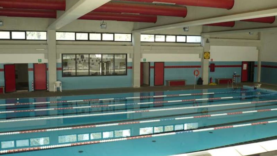Piscina coperta con più corsie delimitate da galleggianti, vasca rettangolare e bordi piastrellati. L’ambiente presenta grandi finestre lungo le pareti, porte rosse e tubazioni rosse a vista sul soffitto con travi bianche.