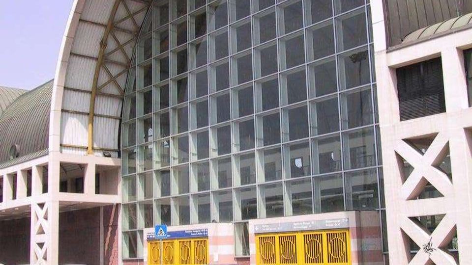 Facciata di un grande edificio pubblico con ampia vetrata a griglia e copertura ad arco in metallo, affiancata da strutture laterali con elementi a X. Davanti si trovano cancelli gialli d’ingresso e un’area stradale con auto parcheggiate.