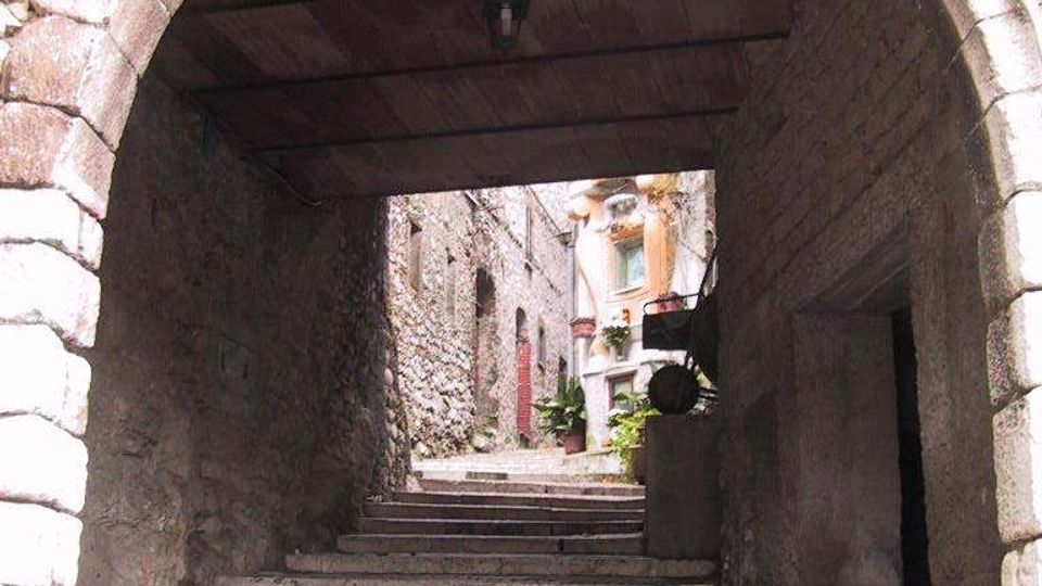 Passaggio ad arco in pietra che copre una scalinata lastricata in un vicolo stretto, con soffitto in legno e pareti in muratura grezza. In fondo si intravedono case in pietra con finestre e piante in vaso lungo i gradini.