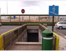Ingresso a un sottopassaggio pedonale sotto una strada, con ringhiere gialle e scala che scende in un vano in cemento. Accanto ci sono un cestino verde e segnali stradali, con barriera antirumore trasparente sullo sfondo e auto sulla carreggiata.