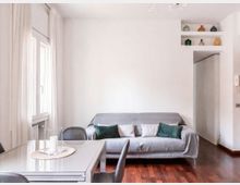 Soggiorno moderno e luminoso con divano grigio e cuscini verdi e chiari, pavimento in legno scuro e tavolo da pranzo con sedie davanti a una grande finestra con tende bianche. A destra si vede un passaggio verso un corridoio e una nicchia a mensole con piccoli vasi decorativi.