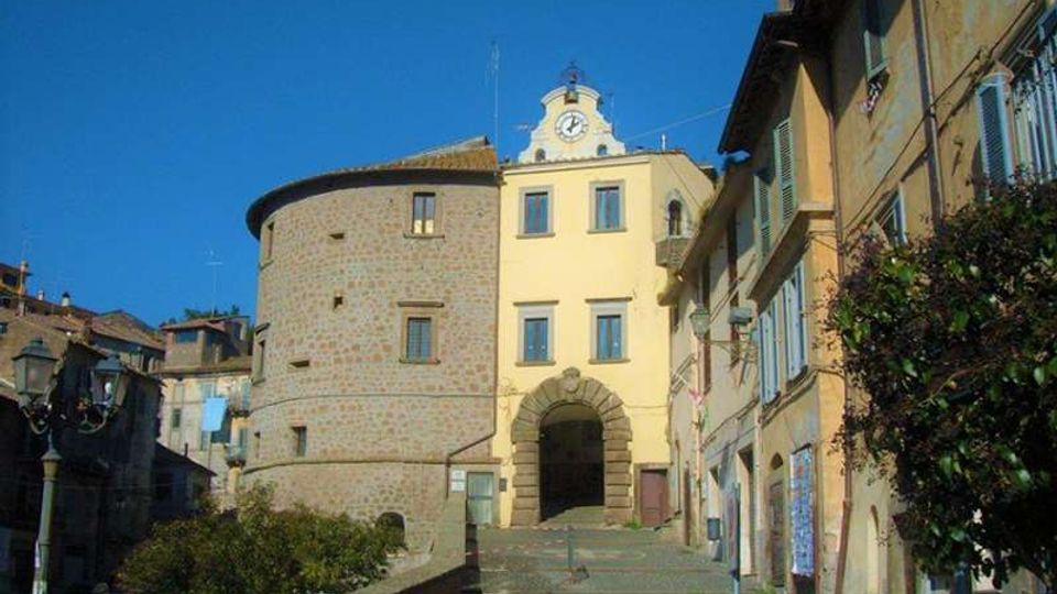 Veduta di una piazza in un borgo storico con edificio giallo a più piani e grande arco d’ingresso in pietra, affiancato da una torre cilindrica in muratura. Ai lati si vedono case con persiane e balconi fioriti, pavimentazione in pietra e lampioni, sotto un cielo sereno.