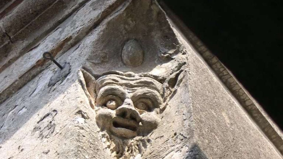 Dettaglio dal basso dell’angolo di un edificio in pietra, con una piccola scultura in rilievo raffigurante una testa grottesca incassata nella muratura. In alto si vede la gronda con tegole e una porzione di cielo.