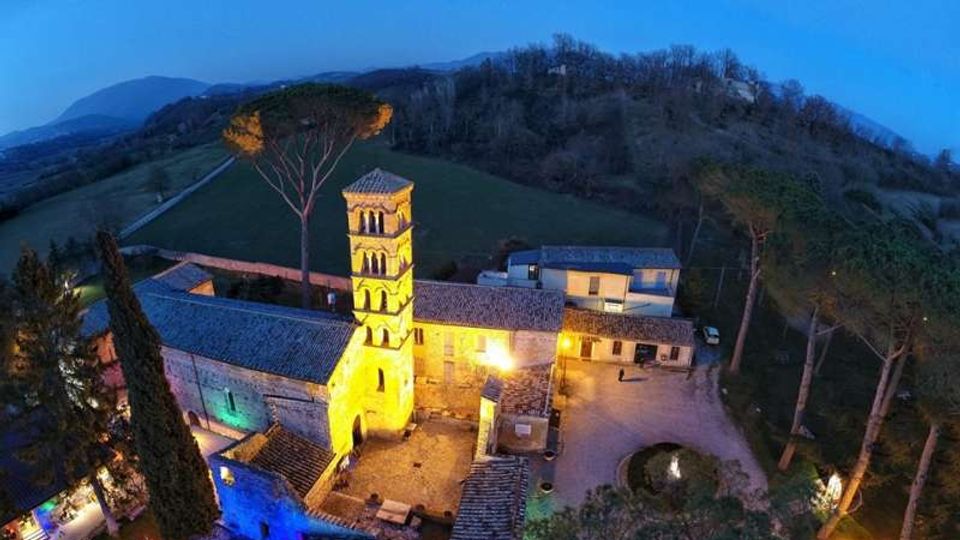 Veduta aerea al crepuscolo di un complesso rurale in pietra con campanile di una piccola chiesa illuminata da luci calde, tetti in coppi e cortile interno. Intorno si vedono colline boscose e campi, con alti pini e un viale che conduce alla struttura.