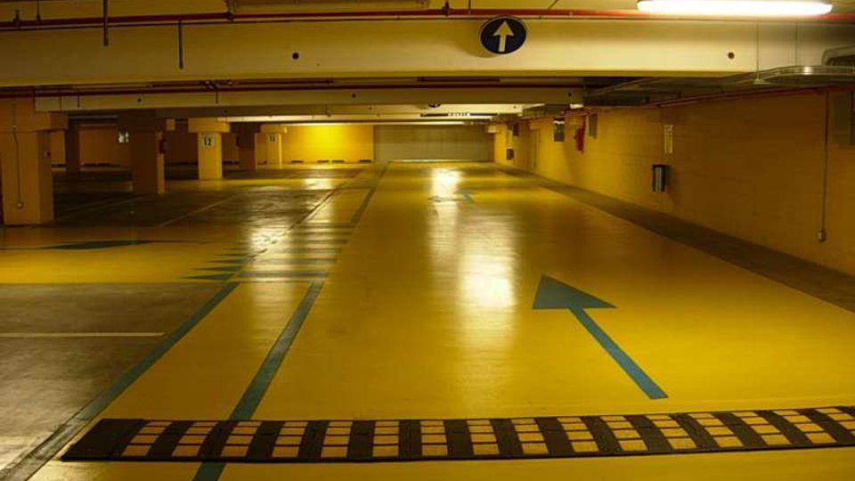 Parcheggio sotterraneo con pavimento giallo e linee blu di corsia, con una grande freccia direzionale dipinta a terra e un dosso rallentatore in primo piano. Soffitto basso con tubazioni e illuminazione al neon, colonne e pareti gialle lungo le corsie.