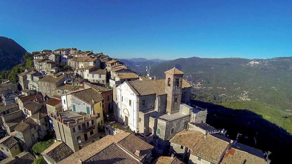 Veduta aerea di un borgo collinare con case in pietra dai tetti in tegole addossate tra loro e una chiesa con campanile al centro. Sullo sfondo si vedono vallate e montagne verdi sotto un cielo limpido.