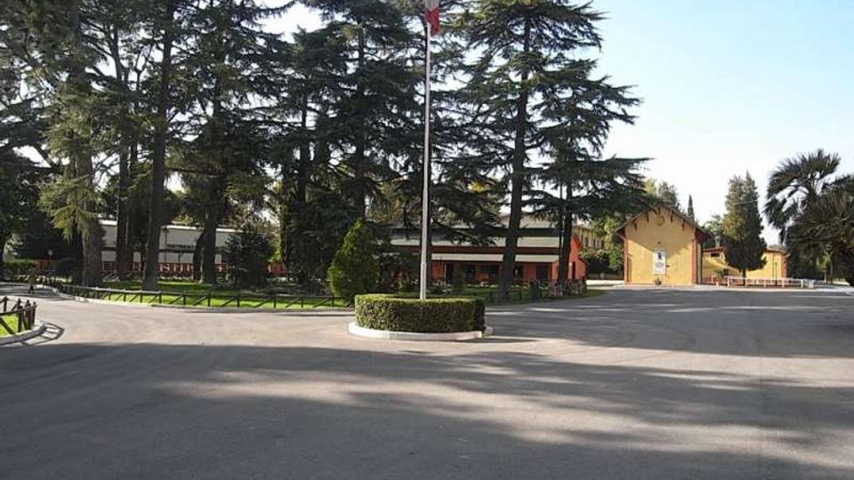 Ampio piazzale asfaltato con aiuola circolare e alto pennone centrale con bandiera. Sullo sfondo si vedono edifici bassi in stile caserma o complesso scolastico, circondati da alti alberi sempreverdi e aree verdi.