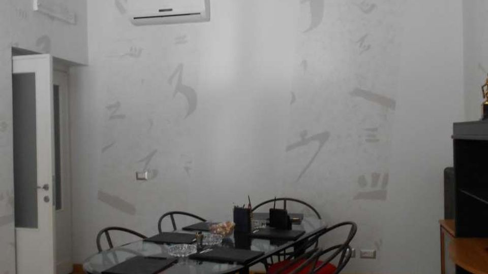 Sala riunioni con pavimento in legno, pareti chiare decorate con segni astratti e climatizzatore a parete. Al centro c’è un tavolo rettangolare con piano in vetro circondato da sedie in metallo con sedute rosse, con faretti a soffitto e una porta a vetri sulla sinistra.