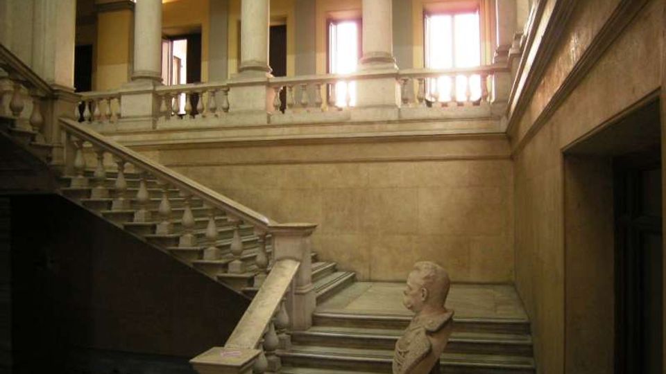 Interno di un edificio storico con ampia scalinata in pietra e balaustra, che conduce a un loggiato con alte colonne e arcate. Grandi finestre sul piano superiore illuminano pareti intonacate e un busto su piedistallo alla base della scala.