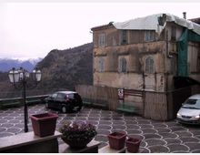 Piazzale panoramico pavimentato con motivi circolari bianchi e neri, con panchine, lampioni e alcune auto parcheggiate. Sul lato destro si vede un edificio in muratura vecchio e deteriorato con impalcatura e teli, sullo sfondo colline e montagne innevate.
