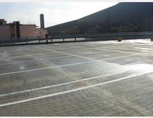 Parcheggio multipiano all’aperto con pavimentazione in cemento e strisce bianche che delimitano gli stalli, circondato da ringhiere metalliche. Sullo sfondo si vedono edifici industriali e una torre verticale sotto un cielo chiaro.