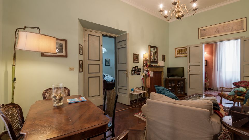 Interno di un soggiorno-sala da pranzo con pareti verde chiaro, pavimento in parquet e lampadario a bracci sul soffitto. Al centro ci sono un tavolo in legno con sedie, un divano chiaro e un’area TV vicino a un camino, con porte a pannelli grigi che conducono ad altre stanze illuminate.